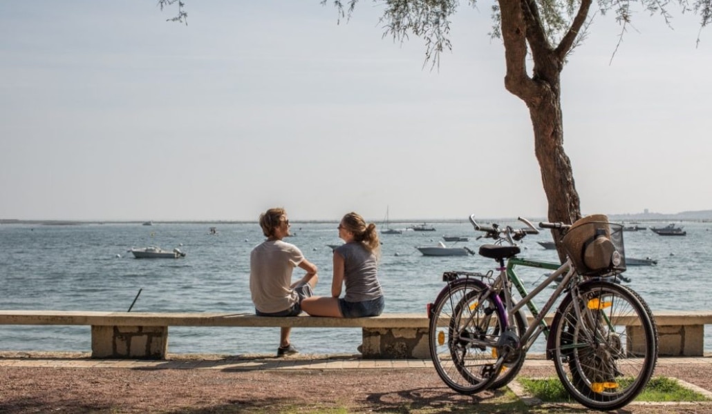 Photo d'un couple assis au bord de la mer avec leurs vélos poséscontre un arbre à coté d'une des pistes cyclables du sud ouest