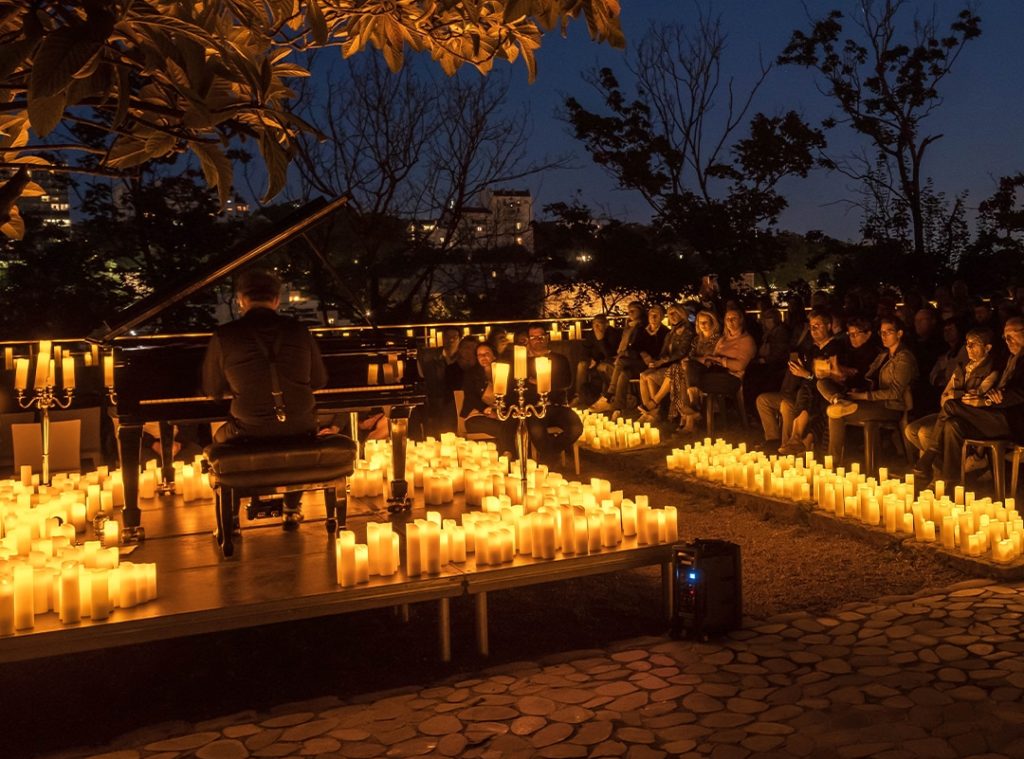 Concert Candlelight en Open Air à Lyon au piano entouré de bougies avec un homme de dos qui joue des morceaux de musique. On voit une partie du public l'écouter