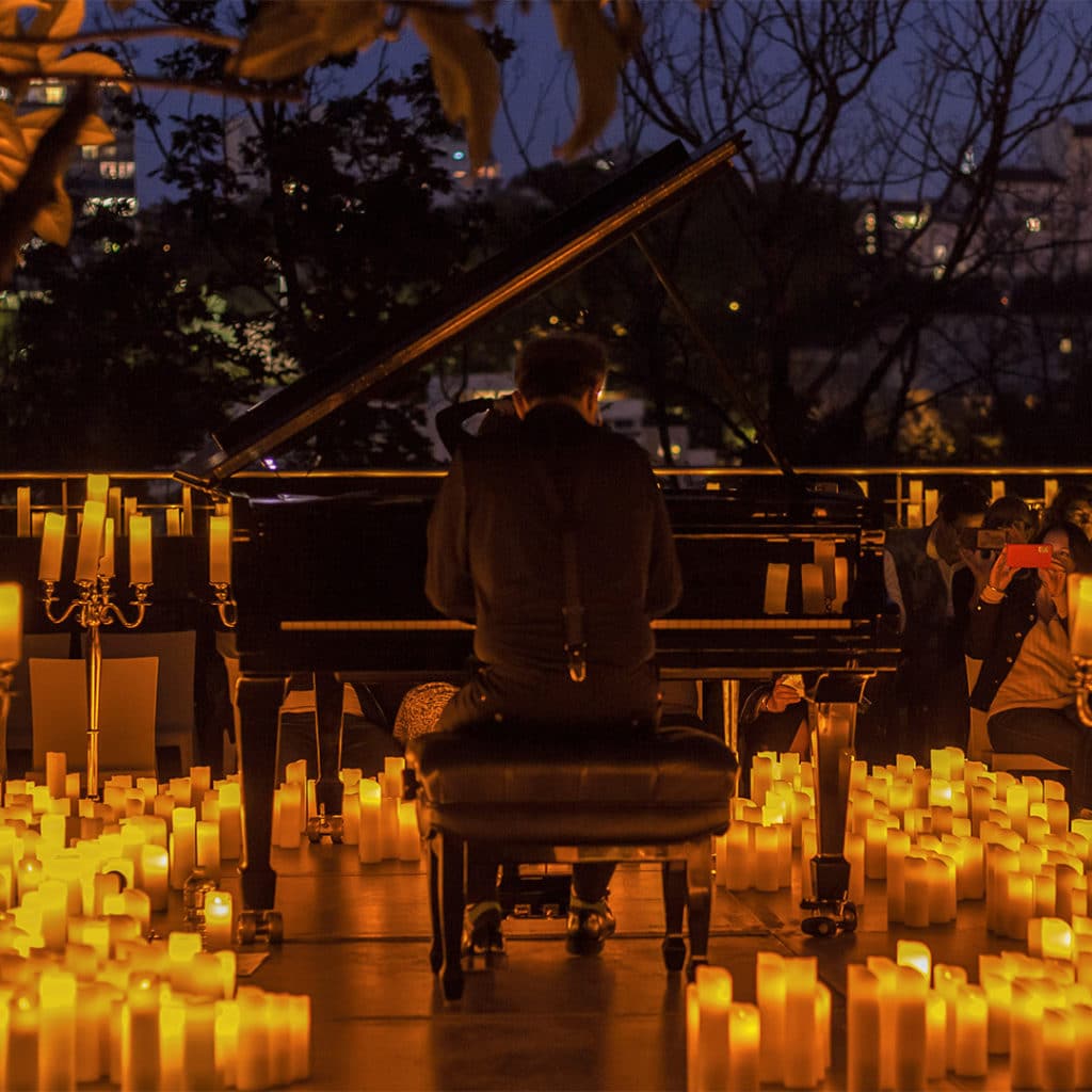 Un pianiste est de dos et joue sur un piano à queue noir avec le sol recouvert de bougies. C'est la nuit, et le pulic en arrière plan prend des photos au téléphone du concert.