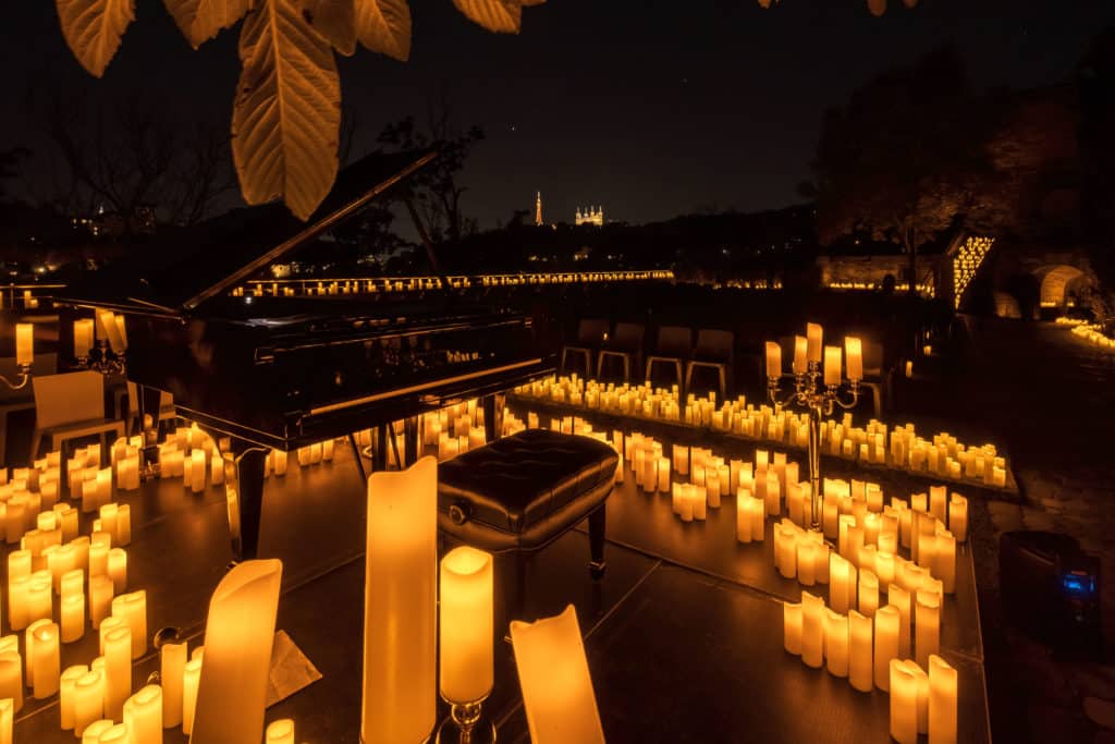 Concert Candlelight à Lyon. On y voit un piano en premier plan éclairé et entouré par des dizaines de bougies en plein air, la nuit.